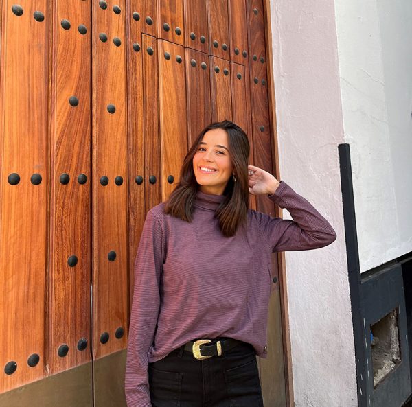 chica delante de una puerta grande marrón con camiseta de rayas moradas y grises con pantalón negro