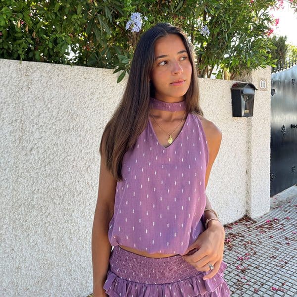 chica en una calle delante de un muro blanco vestida con una blusa cuello halter con escote pico, sin mangas y goma fruncida en cintura en color rosa con detalles de lúrex plateado y falda del mismo tejido y color con cintura fruncida y volantes