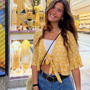chica sonriendo delante de un escaparate con joyas doradas vestida con una blusa amarilla abrochada en frontal con lazada
