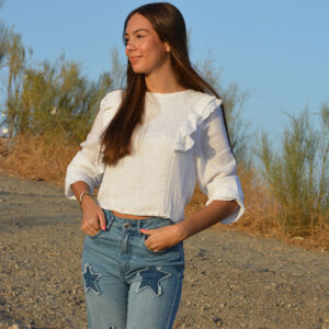 chica en el campo con una blusa blanca con lúrex plata con forro interior blanco y volantes en hombros. Manga larga con goma elástica en puños