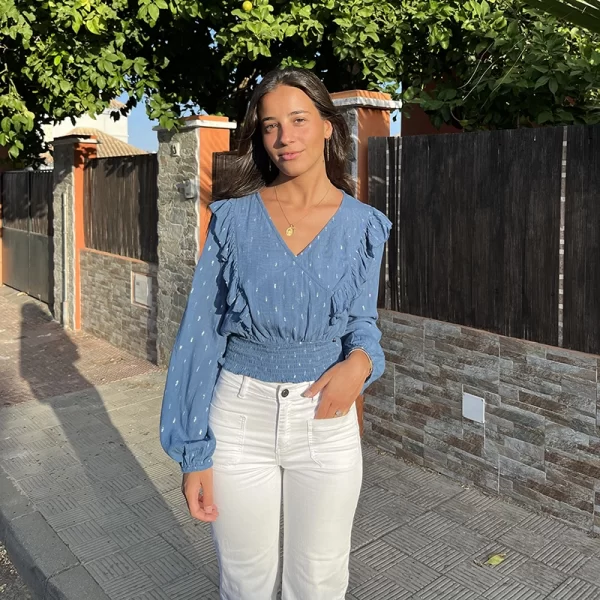 chica sonriendo con la mano en el pantalón vestida con una blusa azul con lúrex plata cruzada y volantes en el frontal