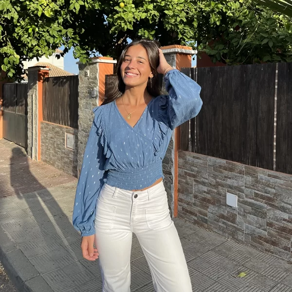 chica sonriendo con la mano en el pelo vestida con una blusa azul con lúrex plata cruzada y volantes en el frontal