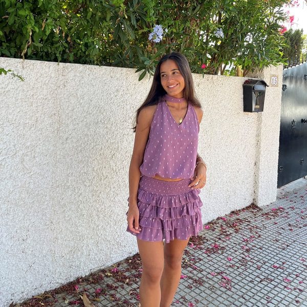 chica en una calle delante de un muro blanco vestida con una blusa cuello halter con escote pico, sin mangas y goma fruncida en cintura en color rosa con detalles de lúrex plateado y falda del mismo tejido y color con cintura fruncida y volantes