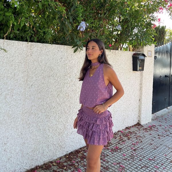 chica en una calle delante de un muro blanco vestida con una blusa cuello halter con escote pico, sin mangas y goma fruncida en cintura en color rosa con detalles de lúrex plateado y falda del mismo tejido y color con cintura fruncida y volantes