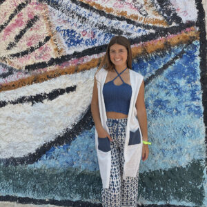 Irene silva delante de una pared llena de grafiti vestido con un pantalón fluido largo en estampado azul y blanco con un top fruncido corto azul y cárdigan blanco sin mangos con bolsillos azules