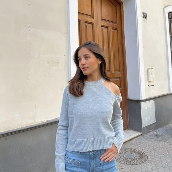 chica delante de una puerta de madera, con una mano en el bolsillo de su pantalón tejano azul con tachuelas y jersey color gris claro con un hombro descubierto