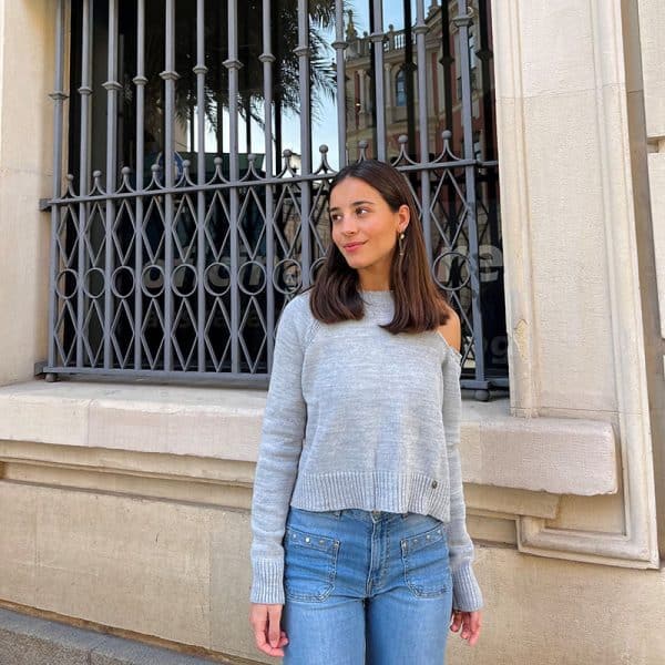 chica delante de una ventana de rejas, con pantalón tejano azul con tachuelas y jersey color gris claro con un hombro descubierto