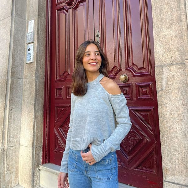 chica delante de una puerta de madera rojiza, con una mano en el bolsillo de su pantalón tejano azul con tachuelas y jersey color gris claro con un hombro descubierto