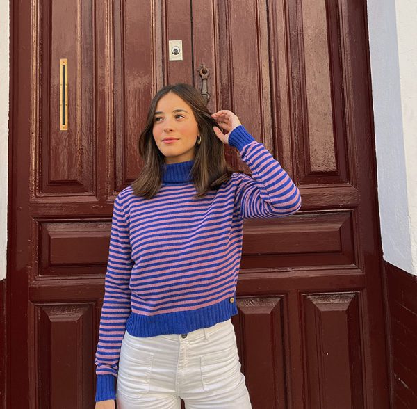 chica sonriendo delante de una puerta grande marrón con un jersey de cuello vuelto azulón en cuello y puños y rayas rosas y azulón en cuerpo