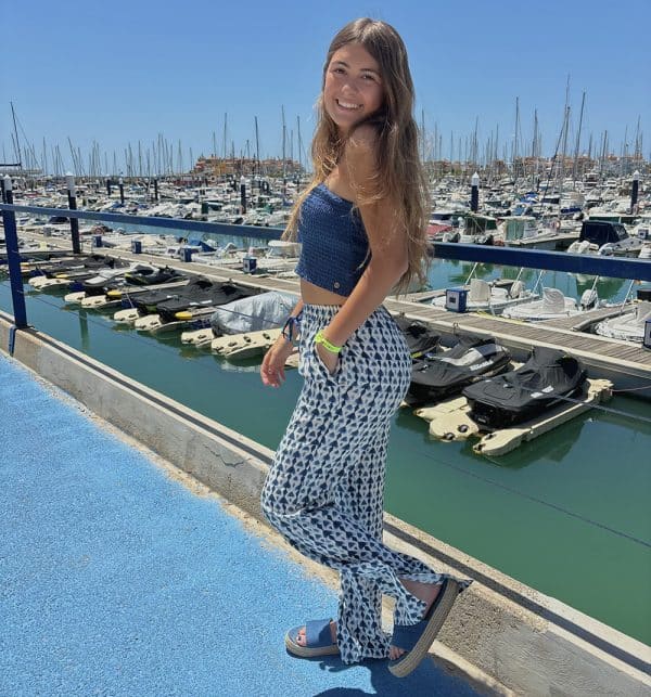 Irene Silva en un puerto lleno de barcos atracados con un pantalón fluido largo estampado azul y blanco con rajas laterales en el bajo y cordón en cintura a tono azul combinado con un top de pink pomelo fruncido con gomas y cuello halter cruzado con cordón en color azul