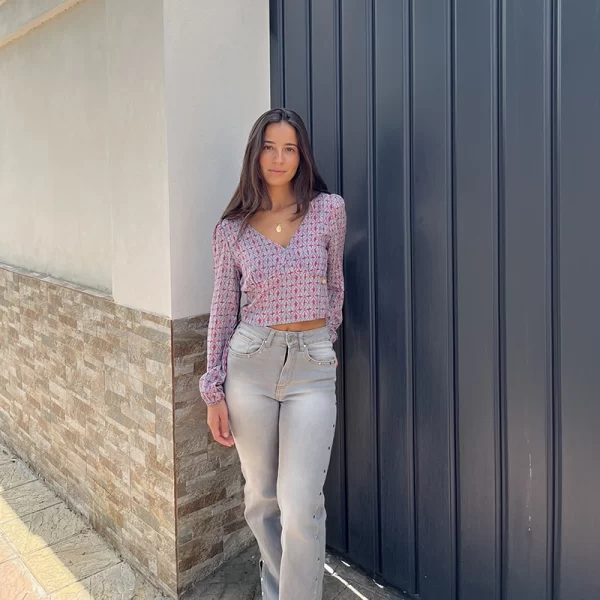 chica en la calle delante de una puerta de garaje gris con blusa modelo Cádiz con estampado verde agua y coral y pantalón gris con tachuelas plateadas