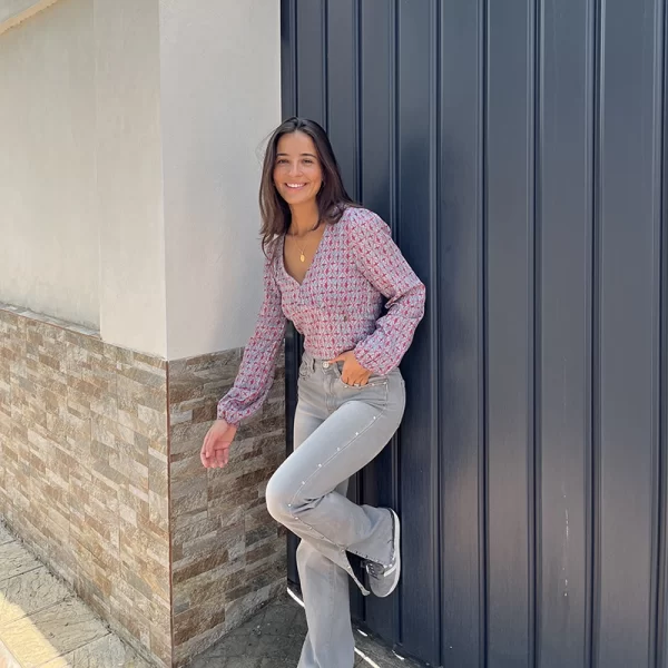 chica sonrtiendo apoyada en una puerta de garaje gris antracita con un pantalón gris con tachuelas plateadas en ambos laterales de las piernas y raja en lateral bajo con una blusa corta escote pico en tonos coral y azul