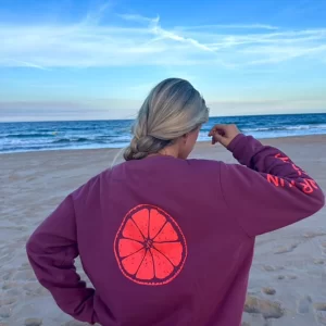 chica en la playa con un pantalón campana gris oscuro y una sudadera de pink pomelo en color burdeos con un estampado grande en espalda de una rodaja de pomelo en color coral flúor