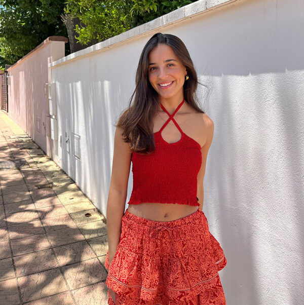 chica sonriendo en la calle delante de una pared blanca de una casa con un top rojo fruncido y atado al cuello y falda corta con dos volantes coral y naranja