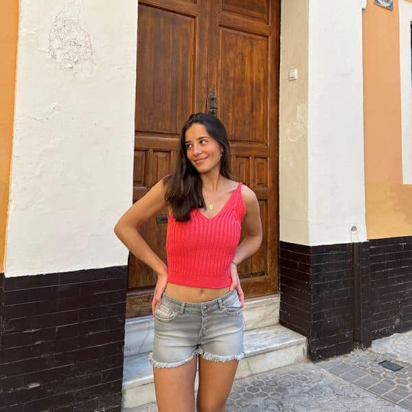 chica delante de una puerta de madera con un top de tricotosa en color coral y un short gris con efecto gastado suave