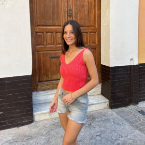 chica delante de una puerta de madera con un top de tricotosa en color coral y un short gris con efecto gastado suave