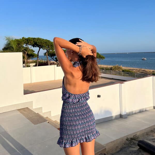 chica posando de espaldas con las manos recogiéndose el pelo en el exterior de una casa con vistas a mar vestida con un vestido azul estampado de cuello halter y parte de la falda fruncida con un volante en el bajo y espada descubierta