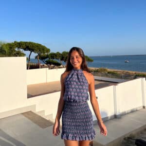 chica posando en el exterior de una casa con vistas a mar vestida con un vestido azul estampado de cuello halter y parte de la falda fruncida con un volante en el bajo